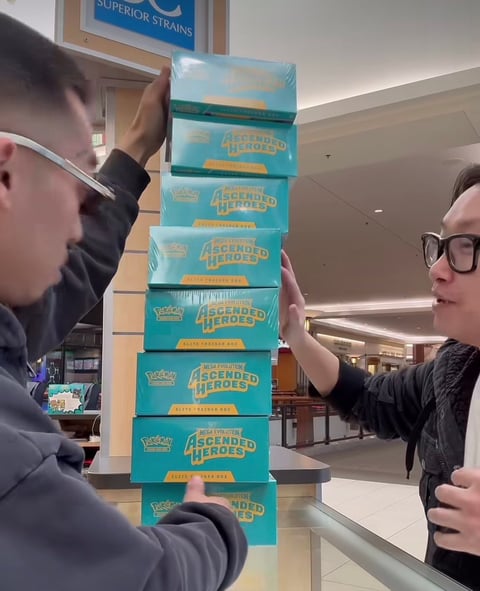 Two people stacking turquoise Pokemon Legends Arceus trading card game booster boxes in a precarious tower indoors